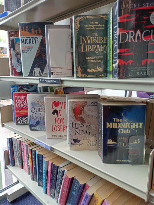 Photograph of a shelf of books with the books angled with the covers facing outwards. Book titles include The Midnight Club by Christopher Pike, The Lies We Sing to the Sea by Sarah Underwood, Love Is For Losers by Wibke Brueggemann, and The Queen of Nothing by Holly Black. 