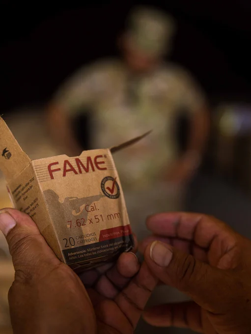 hands holding a small box of ammunition. In the background two individuals dressed in military attire are visible but blurred.