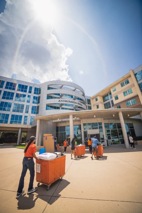 Students moving items using a large orange cart in front of Andromeda Hall on a sunny day with a clear sky and a visible sun halo.