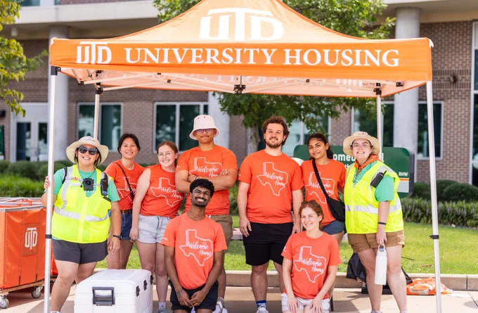 Group of people in orange shirts and hats standing under a University of Texas at Dallas tent outside a building, smiling and posing for the photo.
