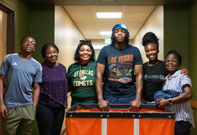 Six individuals standing in a hallway, smiling at the camera. One person is wearing a 'UT Dallas Comets' t-shirt and another a 'Las Vegas Racing' t-shirt.