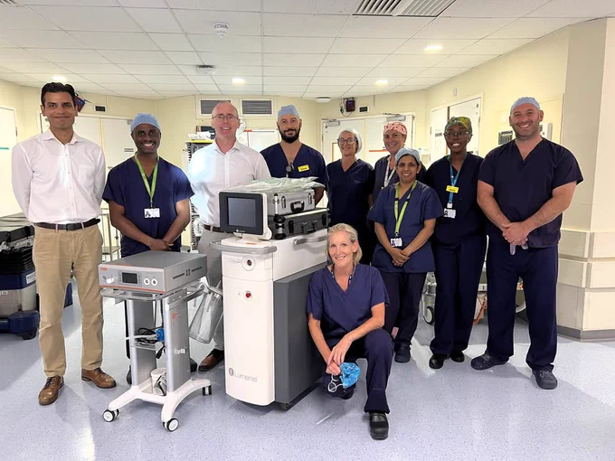 A medical team consisting of nine professionals smiling around advanced hospital equipment in a bright room.