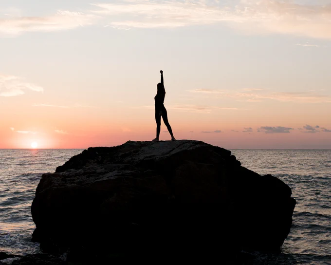 This image shows a silhouetted person standing on a seaside rock at sunset with their arm raised.