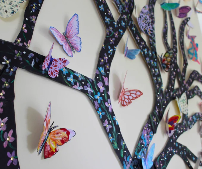 Close up of tree made from card. The bark of the tree has forget me not flowers painted onto it, and there are lots of butterflies made from card which are all painted different colours.