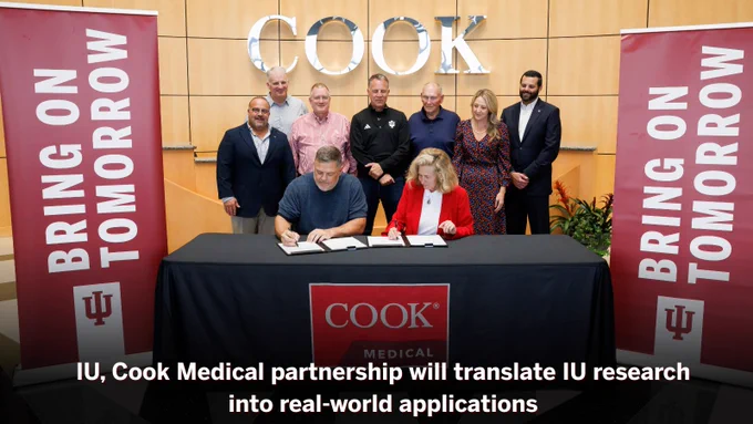 Group of nine individuals gathered around a table at an Indiana University event, where two people are signing a document and others are watching. The backdrop bears the IU and Cook Medical logos with the text "BRING ON TOMORROW".