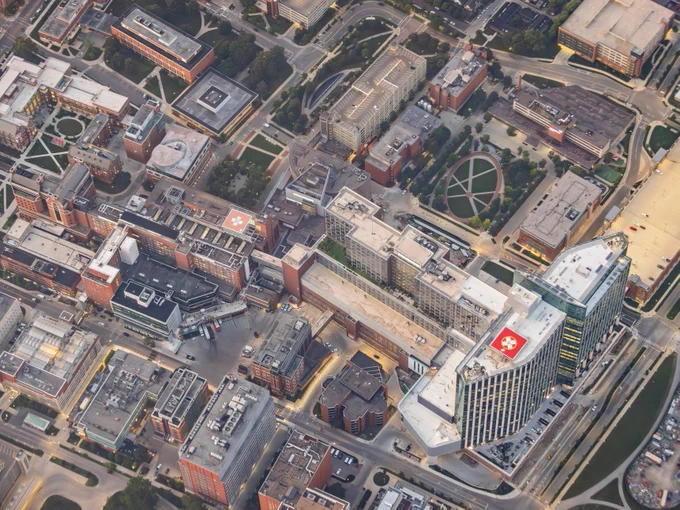 Aerial view of a dense urban area with various buildings, including a prominent hospital marked by a red cross. Streets and smaller structures are interspersed throughout the scene.