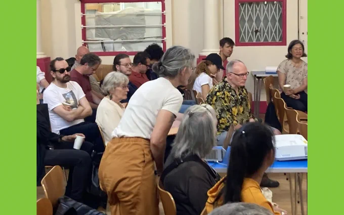 Audience members seated in chairs at a community meeting, attentively listening to a speaker out of view.