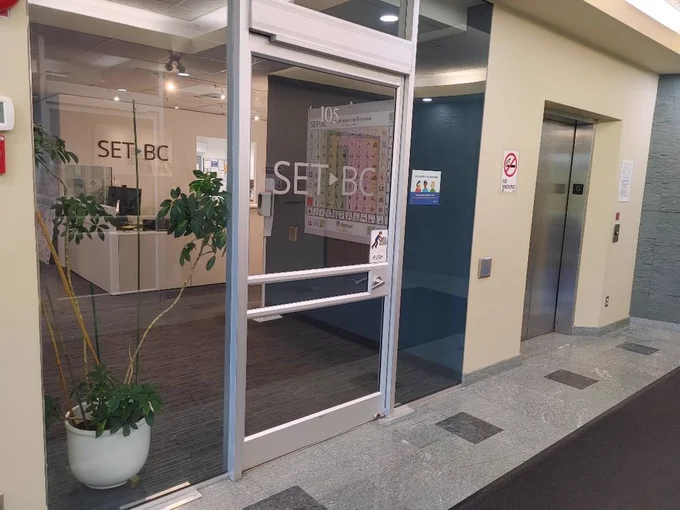 Office entrance with a glass door labeled SET-BC. Inside, desks and an AAC poster are visible. An elevator is to the right.