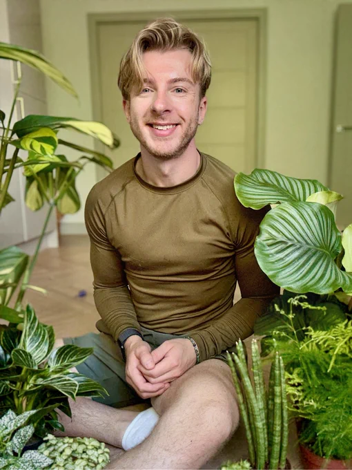 Blonde man smiling sat amongst a selection of bright green houseplants.