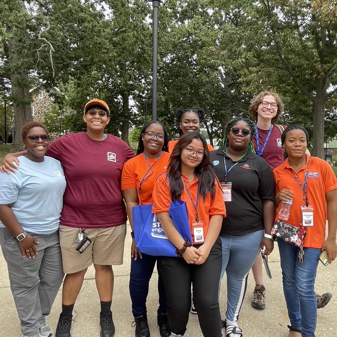 Group of eight students standing outdoors in front of trees and a partially visible building. They wear lanyards with name tags and shirts in maroon, orange, light blue, black, and dark blue. Some hold items like a blue tote bag and a water bottle.