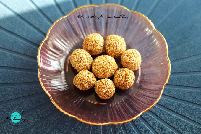 A pink transparent food grade plastic plate (from Temu) with golden sesame seed laddus (Til-Gur Laddu) neatly arranged in the centre, placed on a textured dark blue surface. Captured by blogger Anamika chattopadhyaya for her Everything Enchanting blog and SM pages.