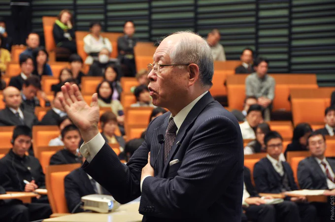Ryoji Noyori in profile giving a lecture to a group of students sitting in the background.