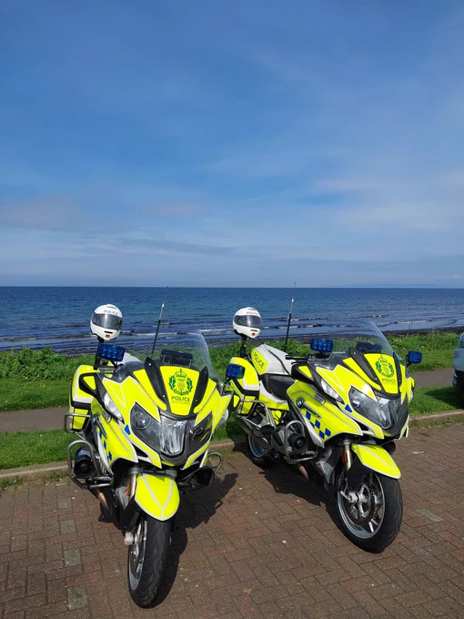 Two marked Police Scotland BMW RT1250 motorcycles parked at the side of the road. There is an expanse of water in the background.