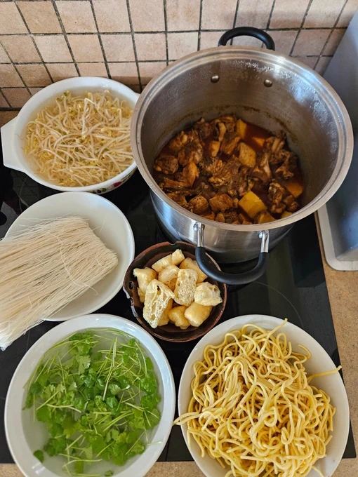 Ingredients cllockwise from top left of a bowl of beansprouts, a pot of chicken & potato curry, a bowl of tofu puffs, a bowl of yellow noodles, a bowl of fresh coriander, a bowl of dry rice vermicelli.