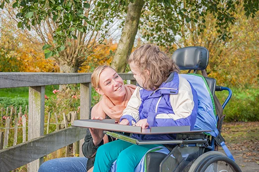A woman and a child in a wheelchair share a joyful moment outdoors, surrounded by autumn trees and a wooden fence, conveying warmth and companionship.