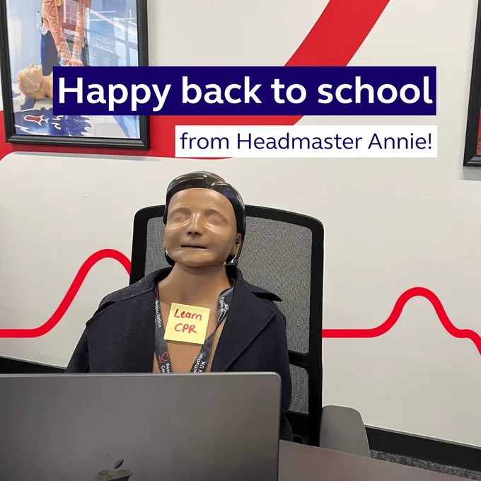 A CPR training mannequin named Annie seated at a desk in a classroom with a laptop, wearing a lanyard labeled "Learn CPR." A banner overhead reads "Happy back to school from Headmaster Annie!"