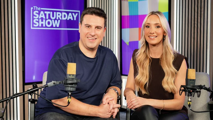 Steven Mill and Amy Irons smile sitting down in front of 2 microphones. In the background is a screen that says The Saturday Show.