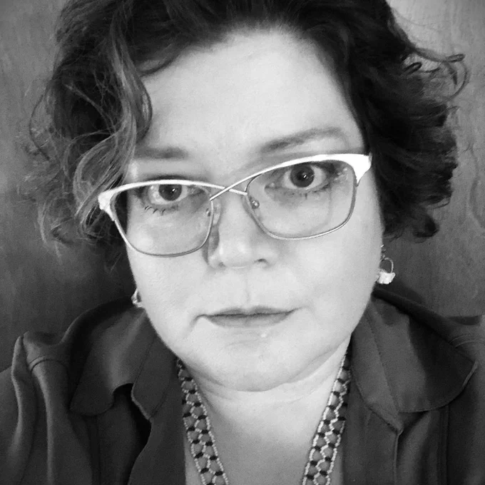 Person wearing a collared shirt and a beaded necklace, with curly hair styled loosely around the head.