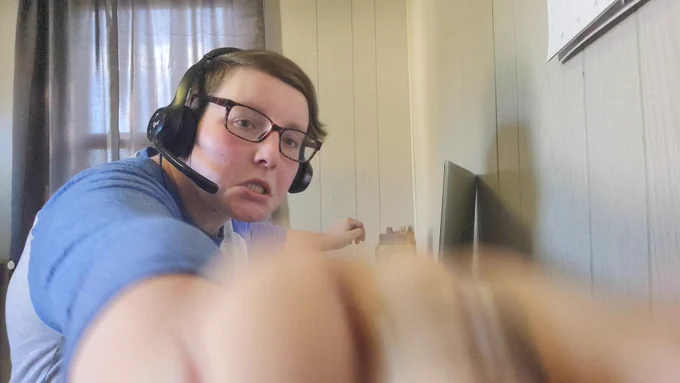 White woman with dark hair and a headset on sitting at a desk with a laptop. Her arm is blurry because outstretched to touch the camera and her face is scrunched in an unflattering but funny expression.