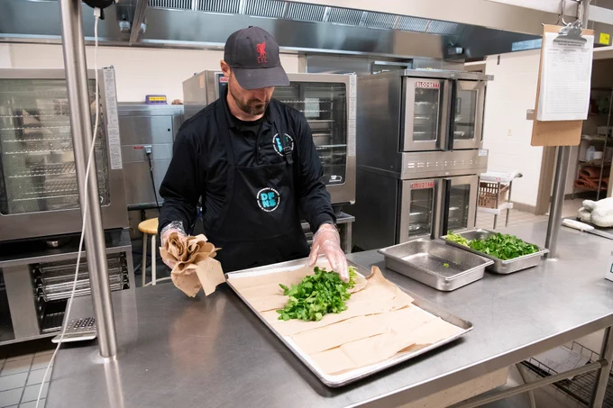 Chef wearing a black apron and cap arranging fresh green herbs on parchment paper in a commercial kitchen.