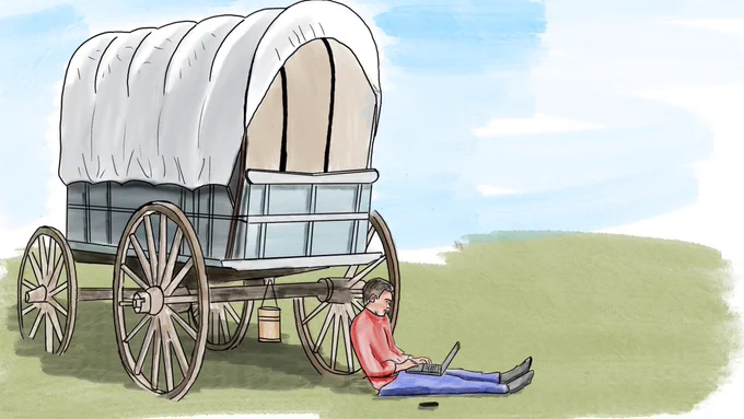A person in a red shirt sits on the ground with a laptop beside a covered wagon with large wooden wheels on a grassy plain under a cloudy sky, blending historical frontier imagery with modern technology