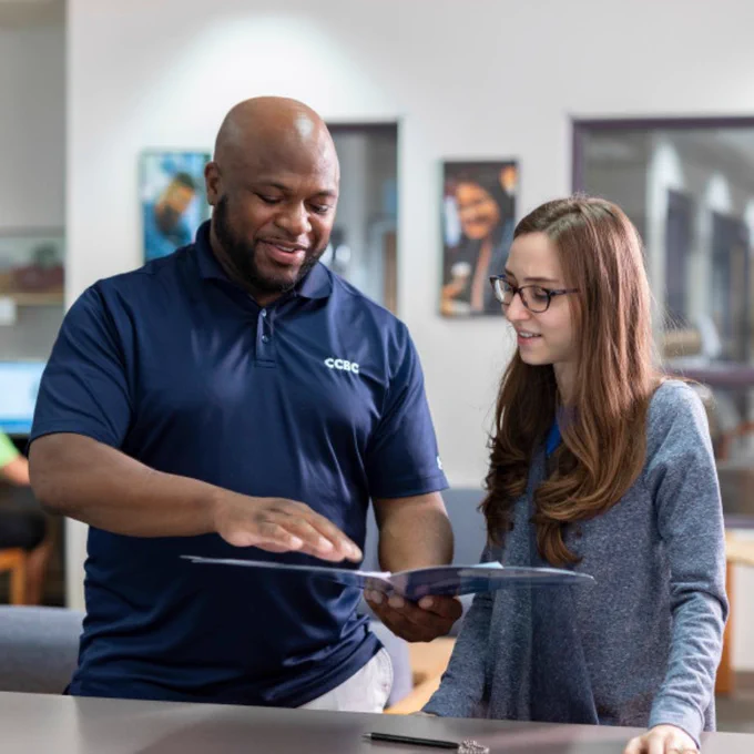 An advisor in a navy blue CCBC polo shirt showing a document to a student in glasses and a gray sweater. They stand at a counter in the Student Services Center, with framed pictures on the wall and another student at a computer in the background.