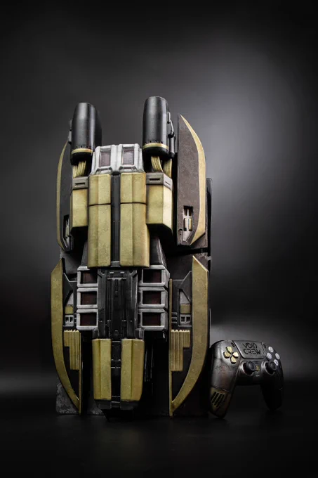 A custom PS5 console and controller against a black background. The console is a custom build with an art deco, sci-fi design and representing a spaceship. It has large, wing-like side plates that are a mix of gunmetal gray and a tarnished, brassy gold. The center section is a dark, sleek gray with detailed lines and panels. To the right of the console is a matching PS5 controller with a similar gunmetal and brassy finish. The right grip of the controller has the words "Void Crew" on it.