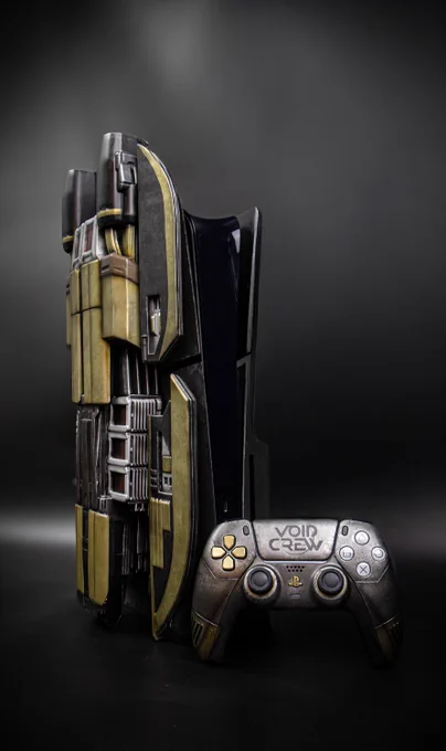 A custom PS5 console and controller against a black background. The console is a custom build with an art deco, sci-fi design and representing a spaceship. It has large, wing-like side plates that are a mix of gunmetal gray and a tarnished, brassy gold. The center section is a dark, sleek gray with detailed lines and panels. To the right of the console is a matching PS5 controller with a similar gunmetal and brassy finish. The right grip of the controller has the words "Void Crew" on it.