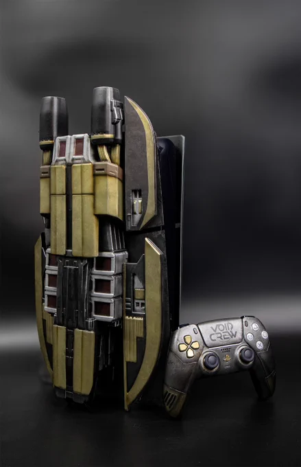 A custom PS5 console and controller against a black background. The console is a custom build with an art deco, sci-fi design and representing a spaceship. It has large, wing-like side plates that are a mix of gunmetal gray and a tarnished, brassy gold. The center section is a dark, sleek gray with detailed lines and panels. To the right of the console is a matching PS5 controller with a similar gunmetal and brassy finish. The right grip of the controller has the words "Void Crew" on it.
