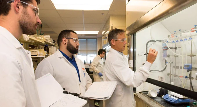 Three individuals in a laboratory observing and taking notes as one person writes on a glass board with diagrams.