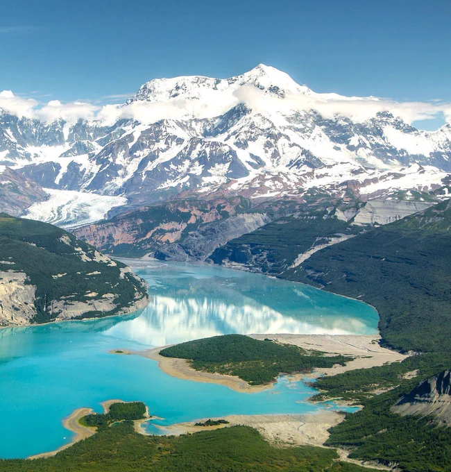 Snowy Mount St. Elias towers above a crystal blue river flowing past the foot of the mountain. 