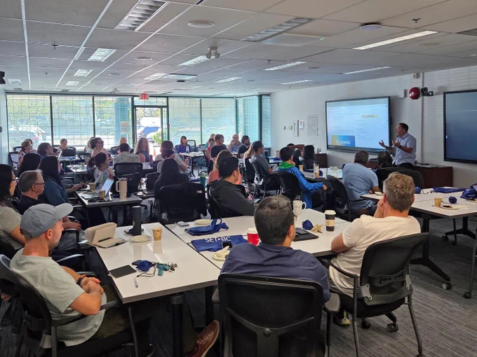A group of people attentively listen to a presentation in a conference room. A man stands at the front, gesturing towards a projected slide.