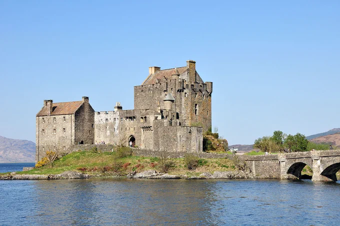 Eilean Donan Castle. The image shows a view over water to the island on which the stone castle stands, with large blocks of the building on the left and right and lower connecting elements between them. On the right is the stone bridge linking the castle with the mainland. More water can be seen on the left. The sky is blue.