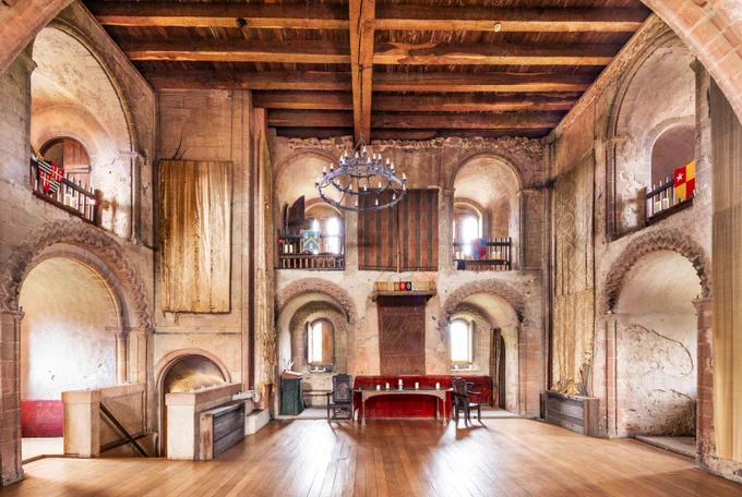 A photograph of the interior of a medieval castle keep with large decorative stone arches around the windows, tapestries hanging on the wall, and an old table and chairs in the centre.