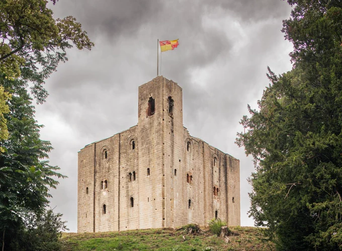 A photograph of the exterior of a medieval castle keep with a flag moving in the wind at the top of a pole at the top of the tower.