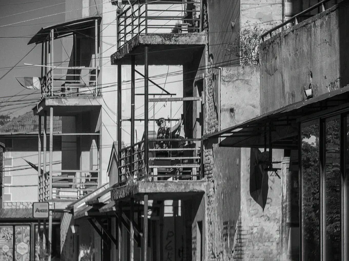 A solitary figure on a balcony, hanging clothes, captured from a distance. The blurred architecture and muted tones transform the scene into something both everyday and poetic — a quiet tension between the ordinary and the abstract. The atmosphere recalls old black-and-white films, evoking memories of forgotten places in Italy, France, or similar cinematic settings.
