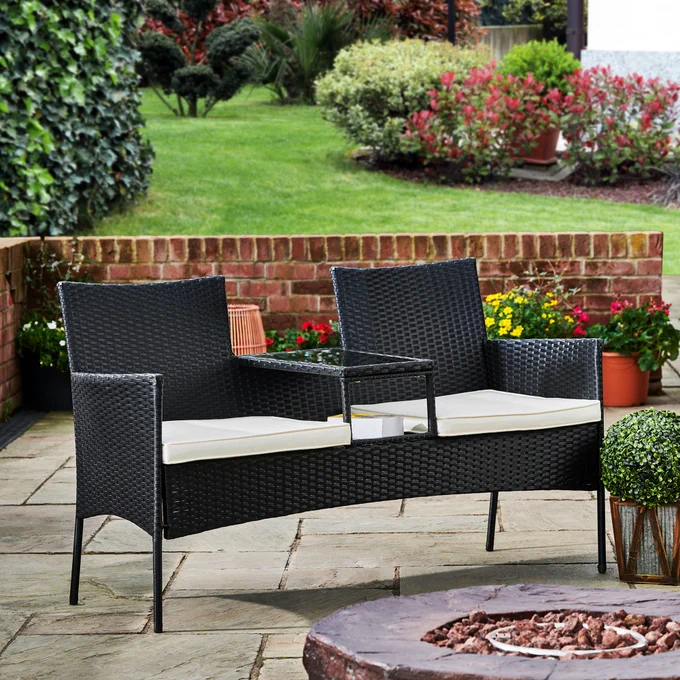 Outdoor patio with two black rattan chairs and a small table, set against a backdrop of vibrant garden flowers and greenery.