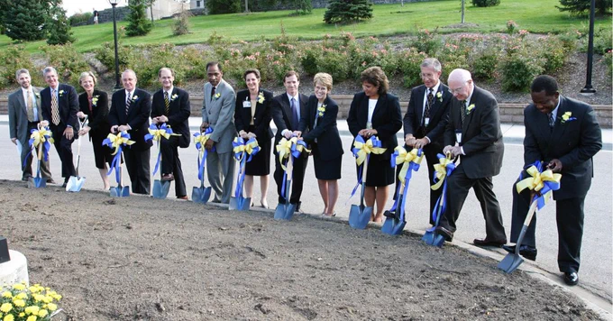 A group of individuals participating in a ceremonial groundbreaking event for the University of Michigan CVC, holding shovels and poised to dig into a patch of soil, with a well-maintained garden and trees in the background.