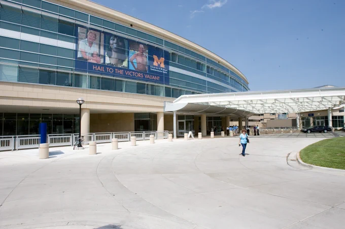Exterior view of the University of Michigan CVC featuring a modern building with large glass windows and a banner that reads "Hail to the Victors Valiant." A few individuals are walking in front of the building.