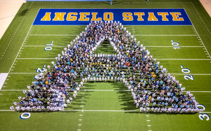 A large group of people arranged to form the letter "A" on a football field, with "Angelo State" displayed prominently in blue and yellow lettering on the end zone. 