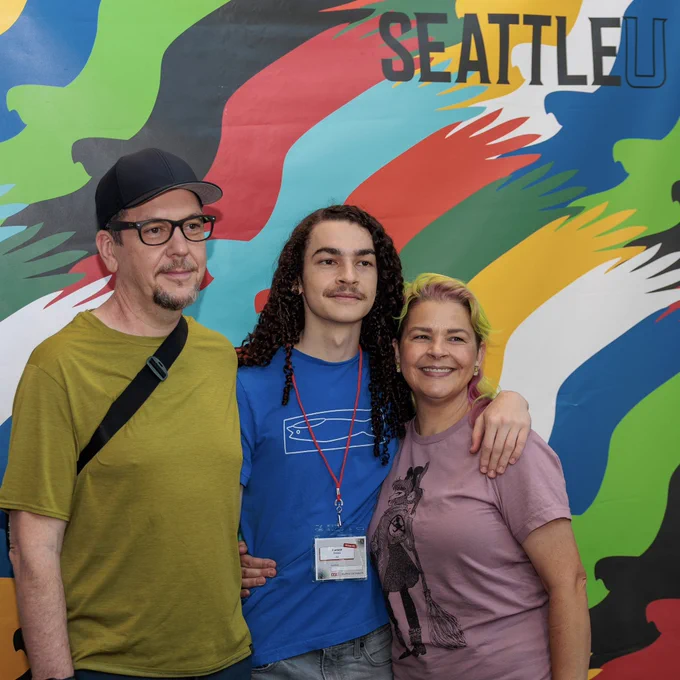 Three people standing and smiling for a photo with a colorful background that says “SeattleU” behind them.