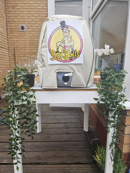 A beer cask with an insulation jacket on, placed on a table draped in flowers. The 'Dry Balls' beer label is affixed to the front of he cask jacket.