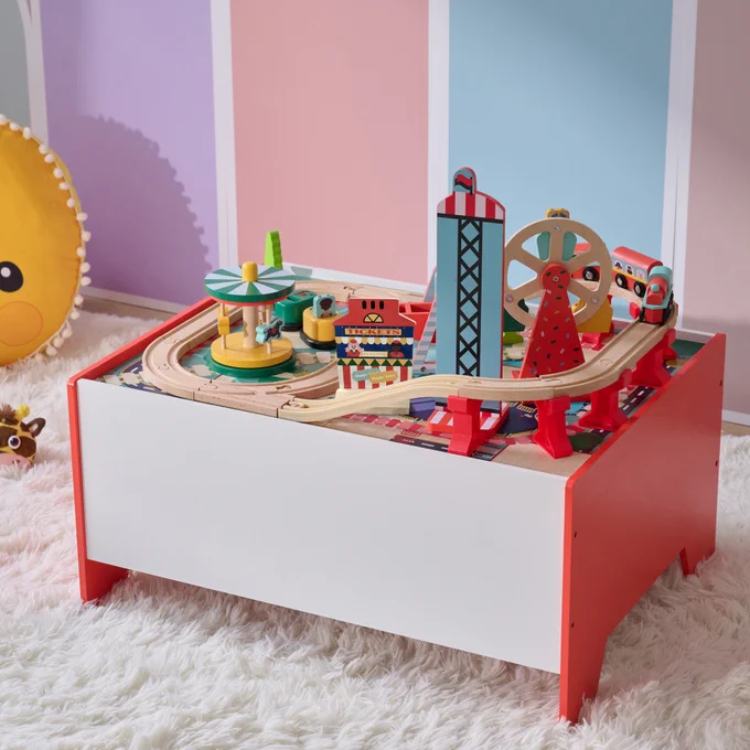 A colorful wooden train set on a table, featuring a railway track, several trains, and various city buildings in a child's playroom.