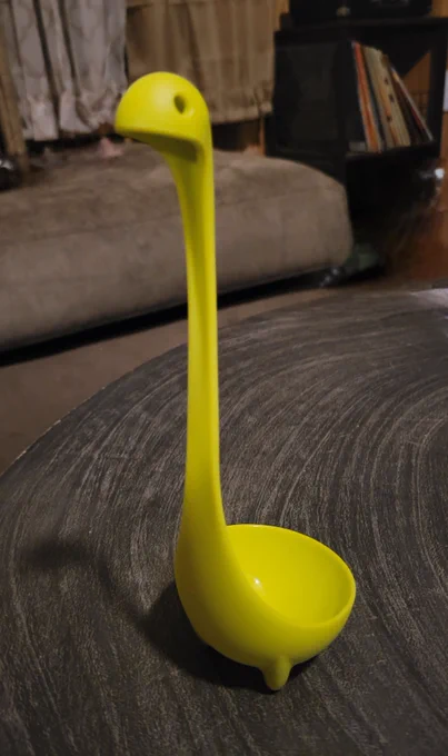 Living room background with dark brown table in foreground. On table is bright neon green hard plastic soup ladle with small triangle feet at the bottom and curves into a small head with small holes for eyes at the top of the handle.