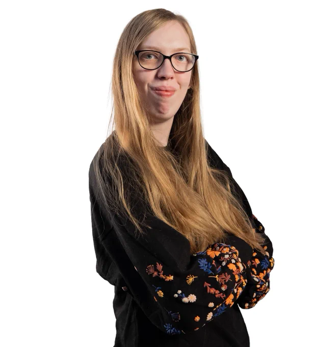 Portrait of a Louella with long blond hair and glasses, arms crossed, wearing a black top with colorful floral sleeves on a white background.