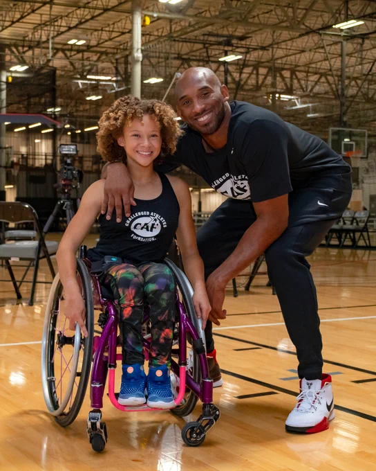 Kobe Bryant squatting with his arm around a young girl in a wheelchair on a basketball court. Both are smiling.