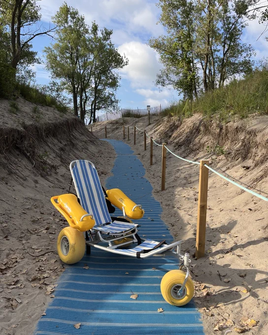A blue and white water wheelchair with large yellow wheels sitting on an accessible beach mat pathway leading onto the beach, with waves crashing in the background under a sky with scattered clouds.