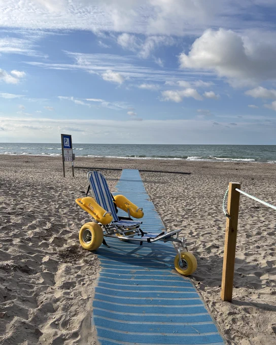A beach wheelchair with large yellow wheels sitting on a beach pathway with blue matting and a roped railing. The pathway leads through the sand dunes into the distance, surrounded by greenery.