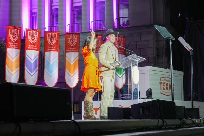 Two individuals at a podium at a University of Texas event, surrounded by banners featuring university shields.
