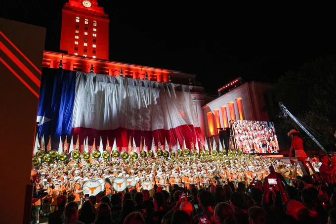 A gathering at the University of Texas, featuring a large band performing in front of the UT Tower, which is illuminated red and decorated with a sizable Texas state flag.